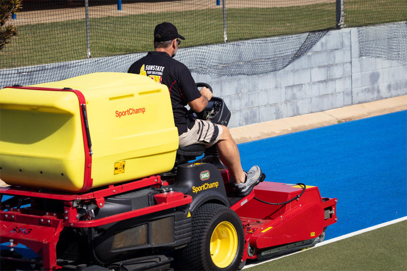 Operator driving the SportChamp machine along the edge of a blue hockey field