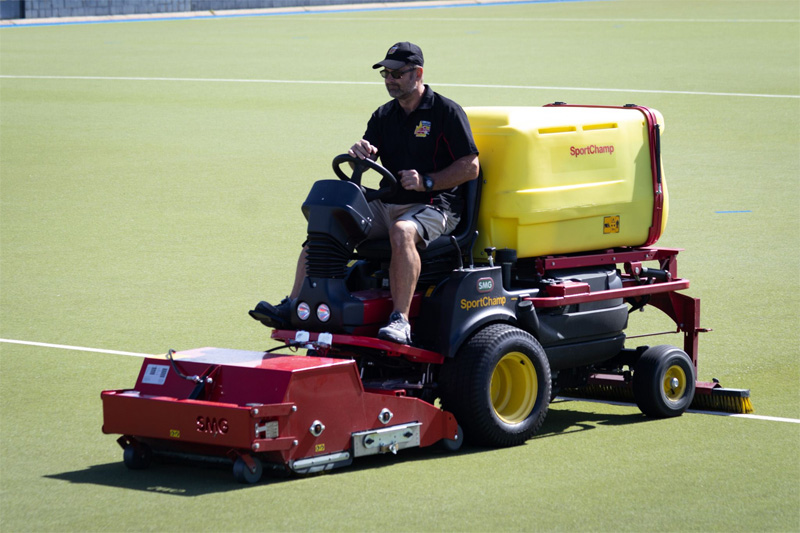 Operator driving the red and yellow turf machine across a green hockey field