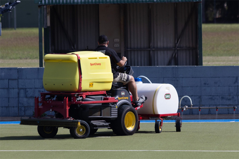 Turf machine towing a white spray tank trailer across a green synthetic field