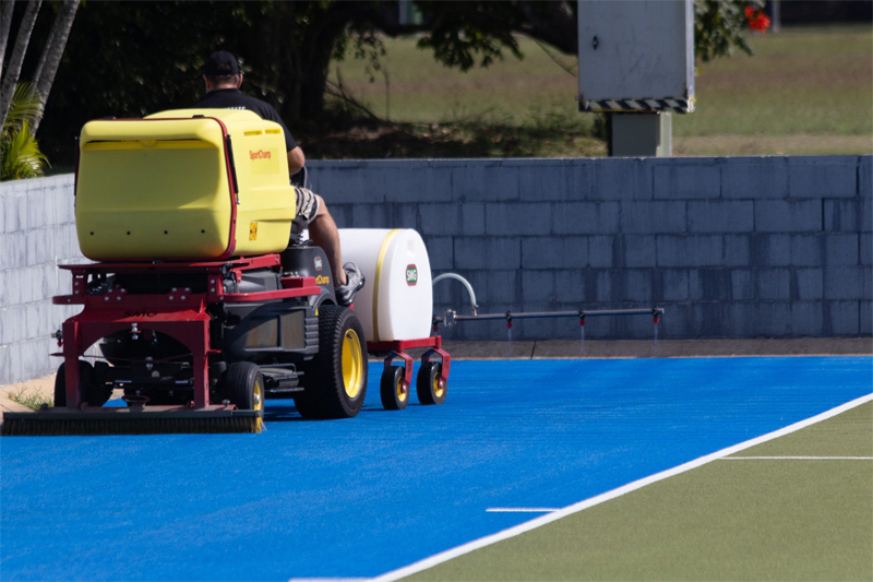 Turf machine towing a spray trailer along a blue synthetic hockey surface