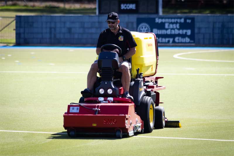 Operator driving a red turf cleaning machine across a synthetic hockey field